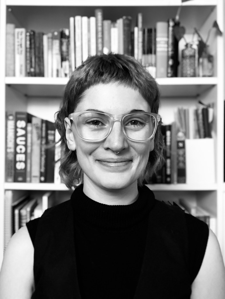 Black and white photo of Nadia sitting in front of a bookshelf. Nadia has a mullet, glasses, and big smile on their face.